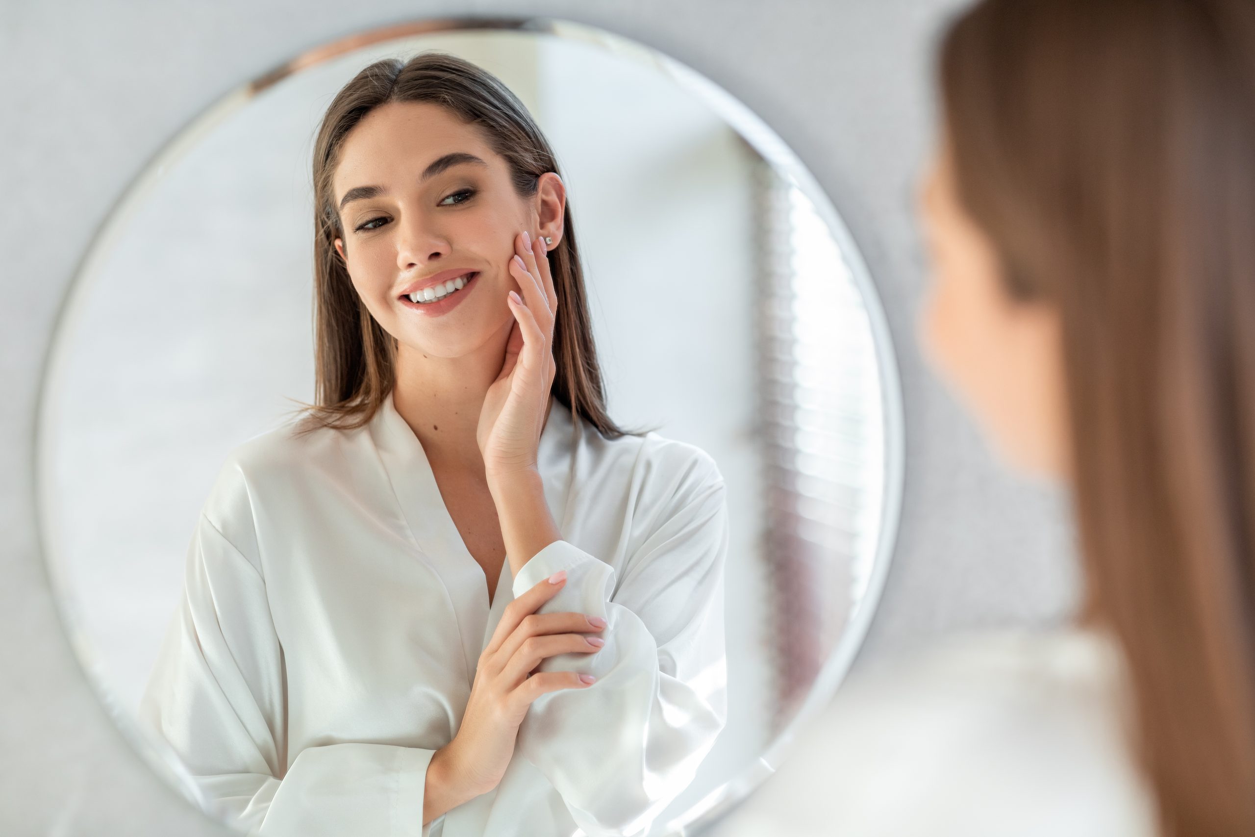 Selbstversorgungskonzept. Portrait von attraktiven jungen weiblichen Blick auf Mirror, schöne Frauen tragen weißes Seidenrobe, die weiche Haut auf Gesicht und Lächeln, genießen ihre Reflexion, selektiver Fokus