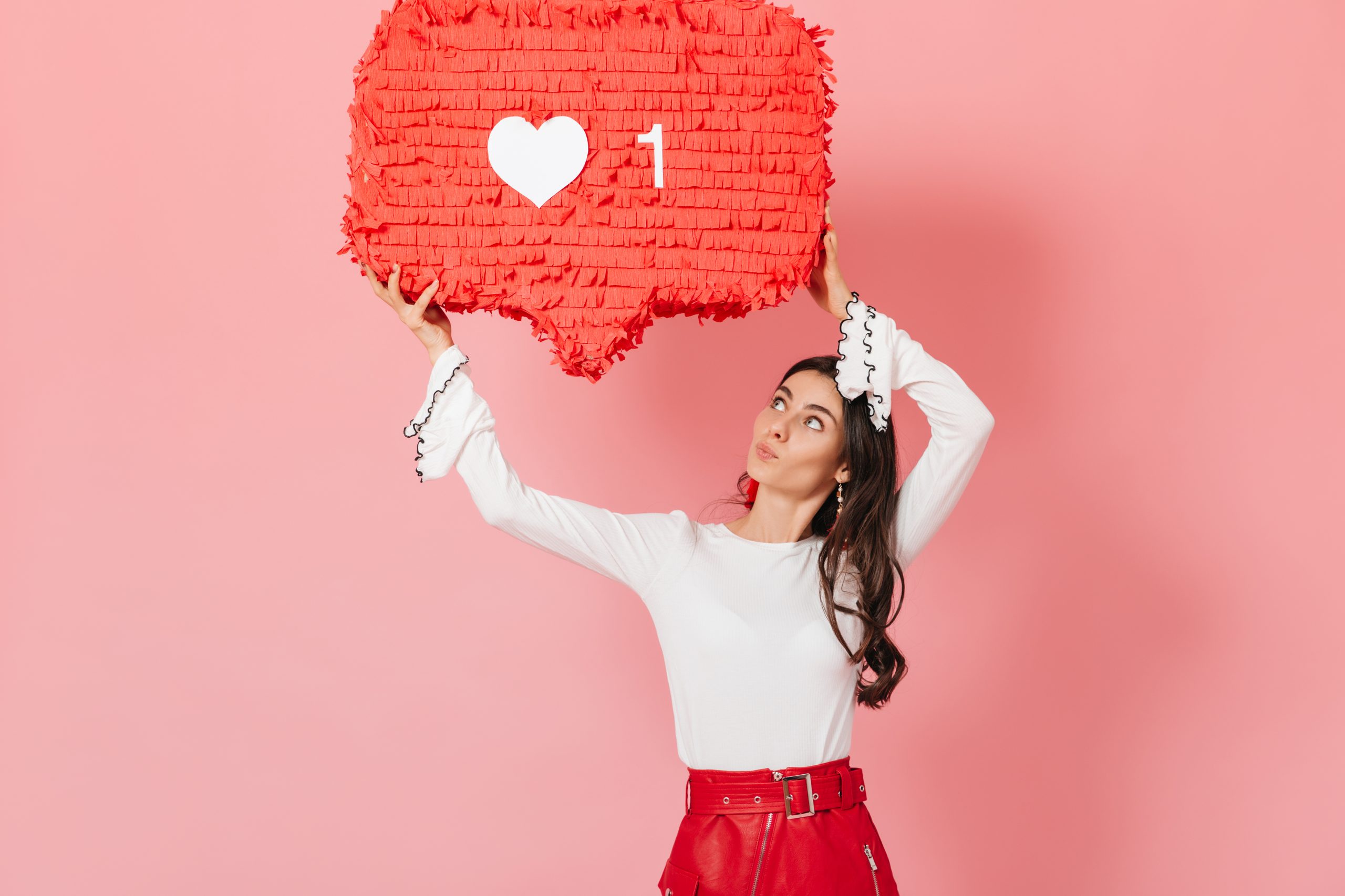 Portrait von Mädchen mit Interesse, die auf ein riesiges "like" Zeichen von Instagram schauen. Brunette auf rotem Rock, auf rosafarbenem Hintergrund