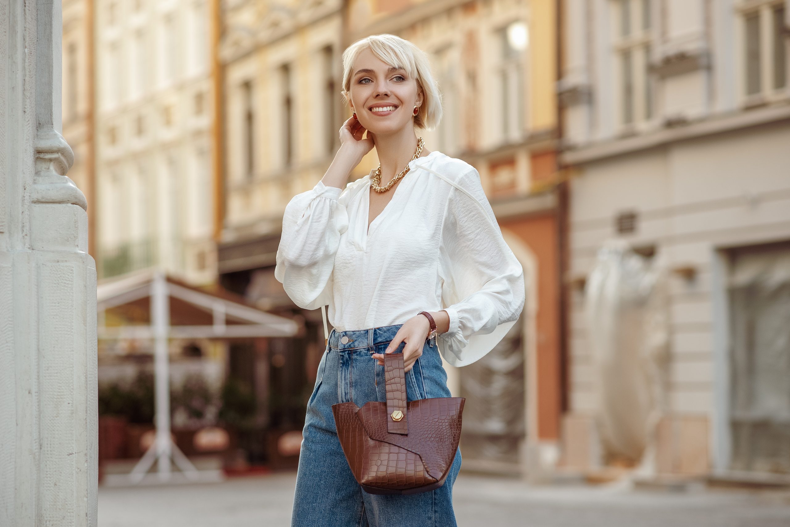 Street-Style Foto von glücklich lächelnden modischen Frau mit trendiger weißer Bluse, High-Tailan Jeans, halten braunen faux Croco-Leder Textur-Tasche. Modell posiert in der Straße der europäischen Stadt.