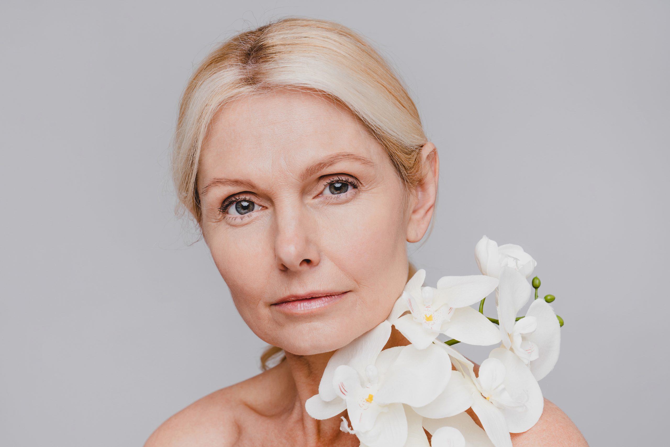 Nahaufnahme eines Porträts einer reifen, hübsch aussehenden Frau mit hübscher Blume nahe ihrem Gesicht einzeln auf grauem Hintergrund