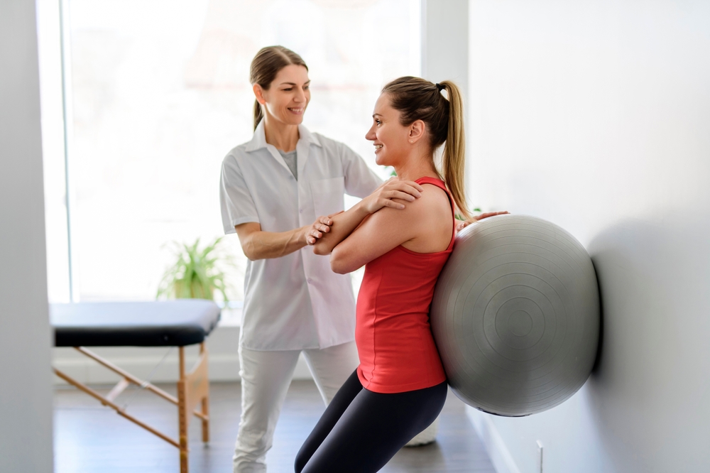 A,Physiotherapist,Helping,Woman,Do,Wall,Squats,With,Fit,Ball.