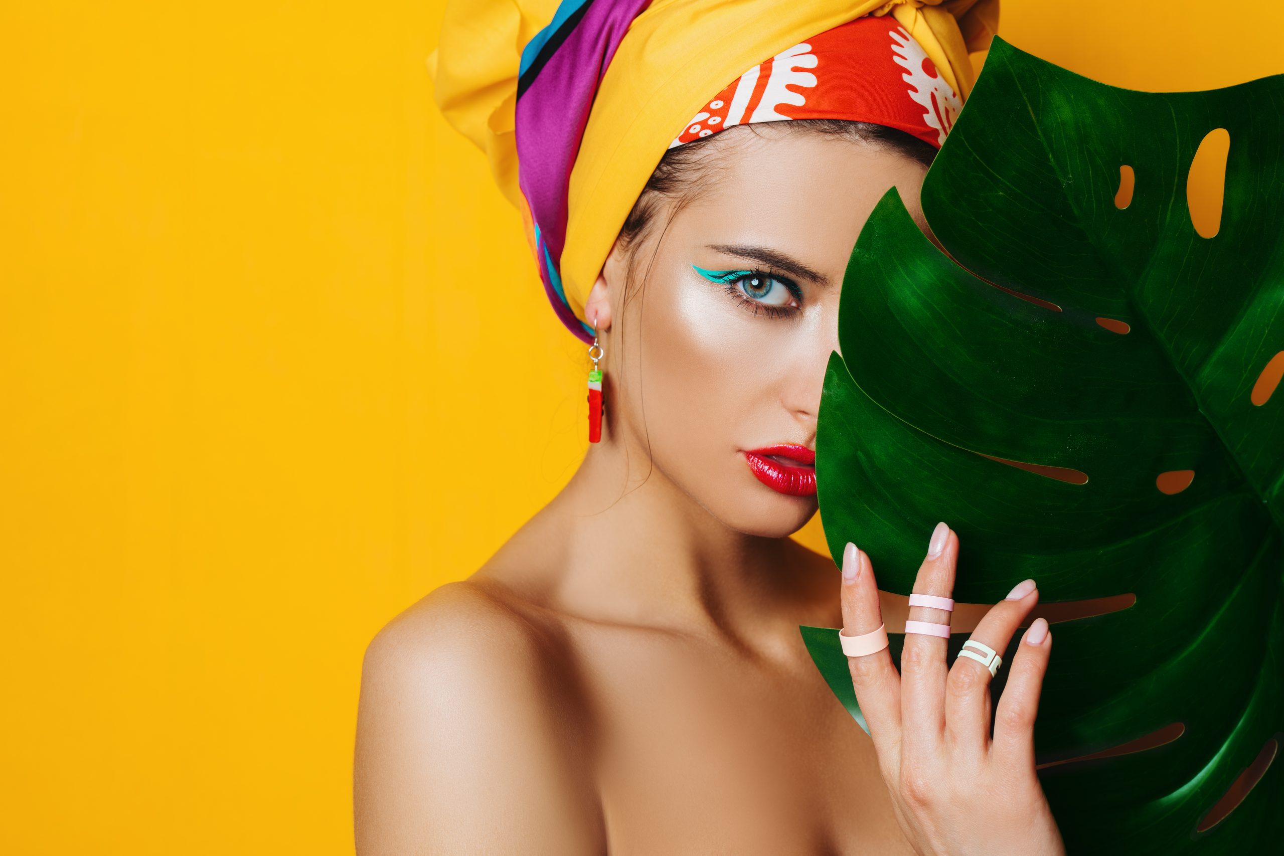 Porträt einer modischen Frau mit heller Schminke hinter einem tropischen Blatt. Gelber Hintergrund. Schönheit, Mode, Make-up-Konzept.