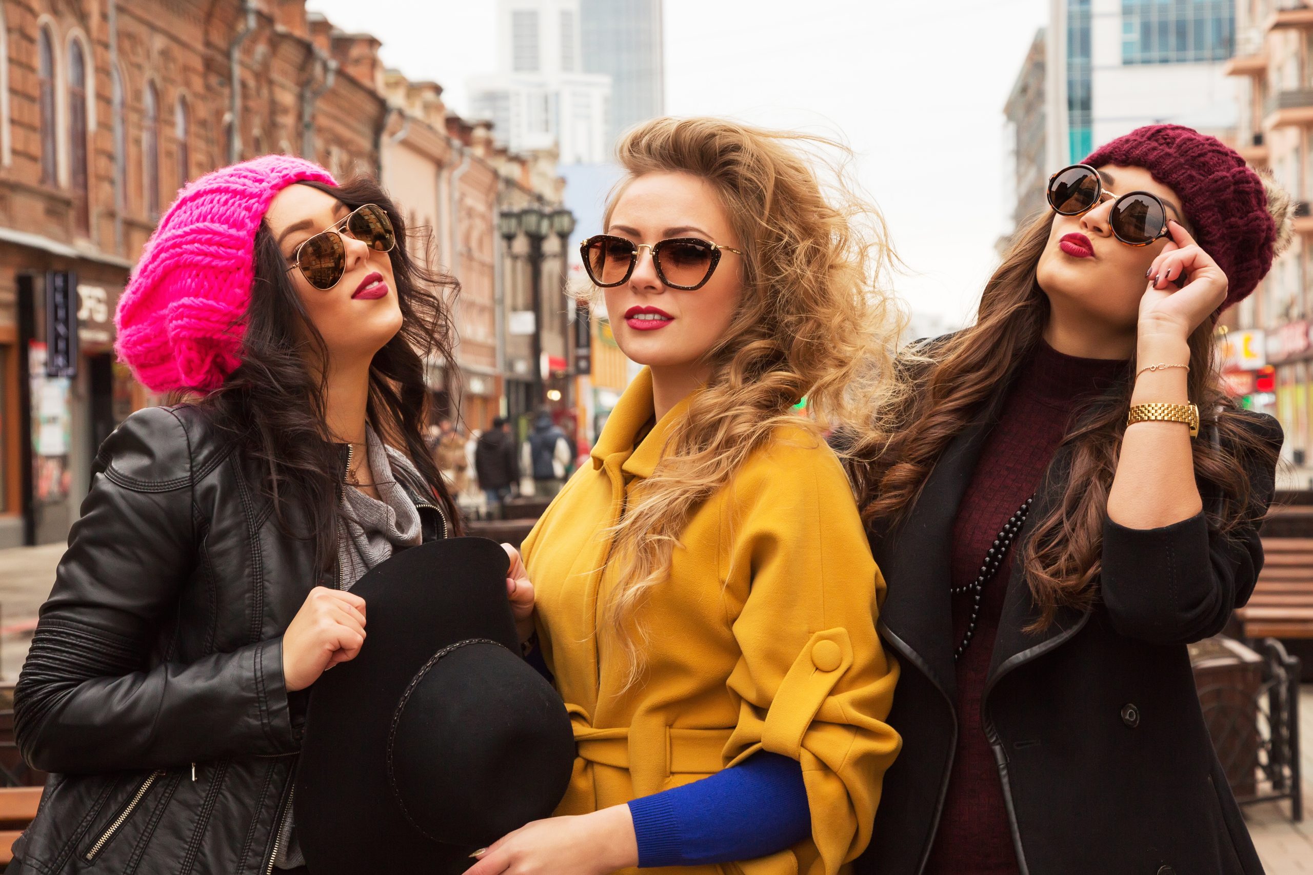 Outdoors fashion portrait of three young pretty smiling girls friends walking at the city. Shopping. Posing at the street. Wearing stylish outerwear, hats and sunglasses. Bright make up. Positive