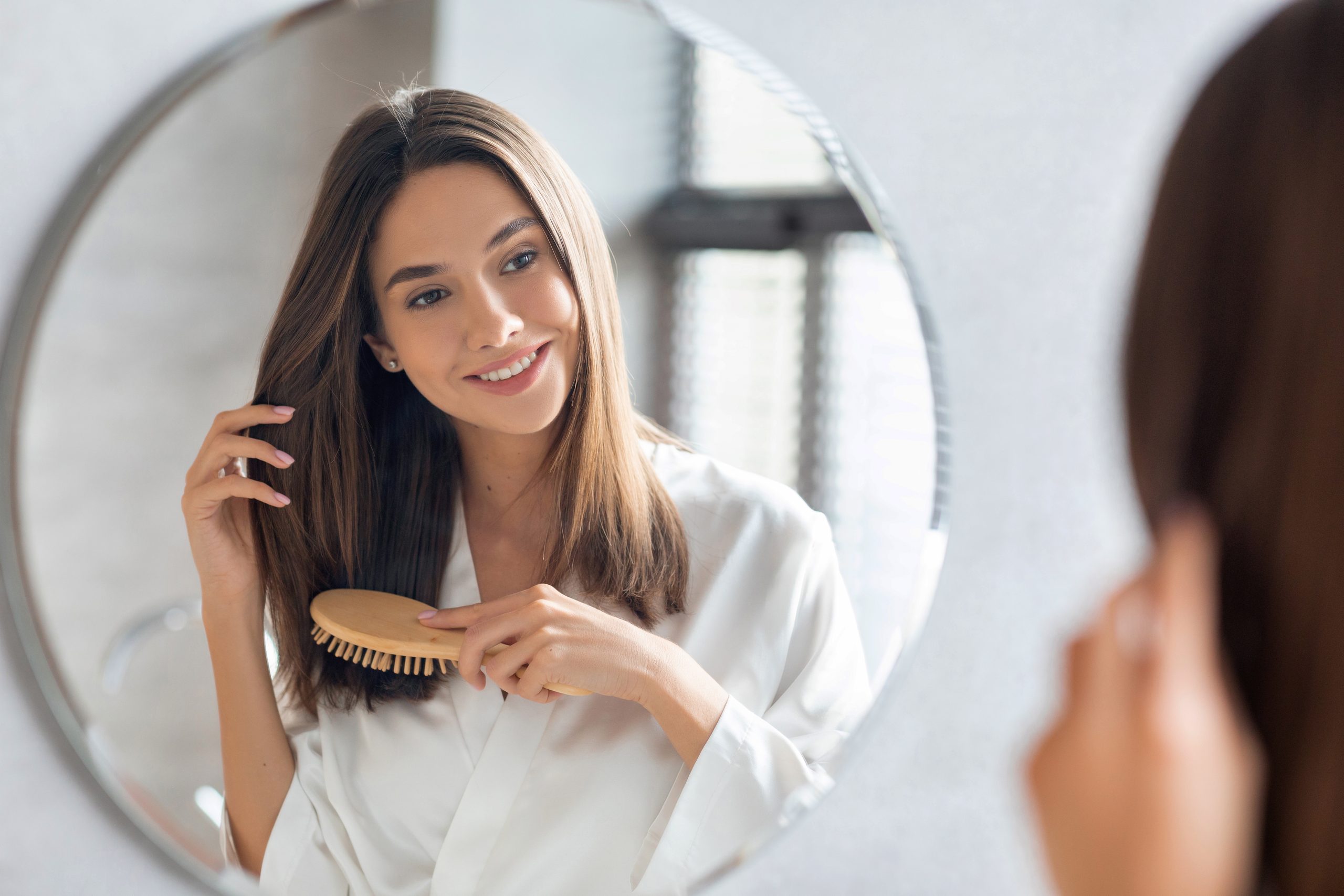 Haarpflegekonzept. Attraktive junge Dame, die ihr dick schönes Haar mit Comb Brush, während sie in der Nähe des Spiegels im Badezimmer steht, glückliche Frau tragen Weiße Seidenrobe Blick auf ihre Reflexion und Lächeln