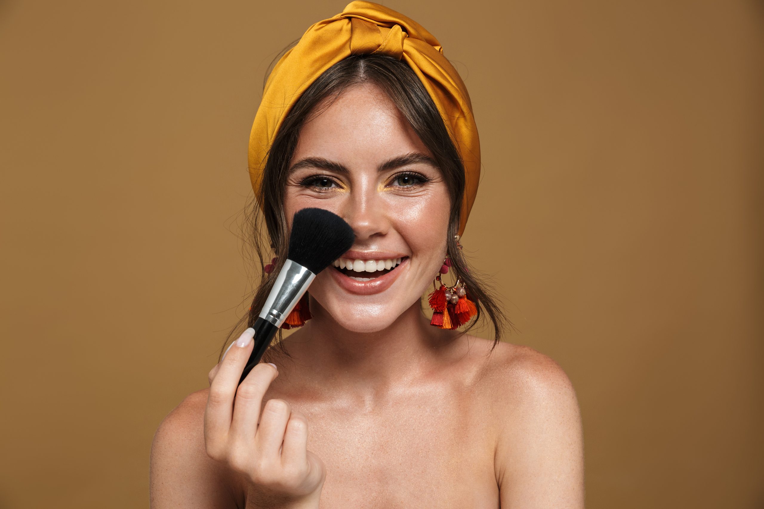 Nahaufnahme eines Kosmetikporträts einer hübschen jungen, toplosen Frau, die ein Kopfband trägt, einzeln auf braunem Hintergrund, mit einer unscharfen Bürste