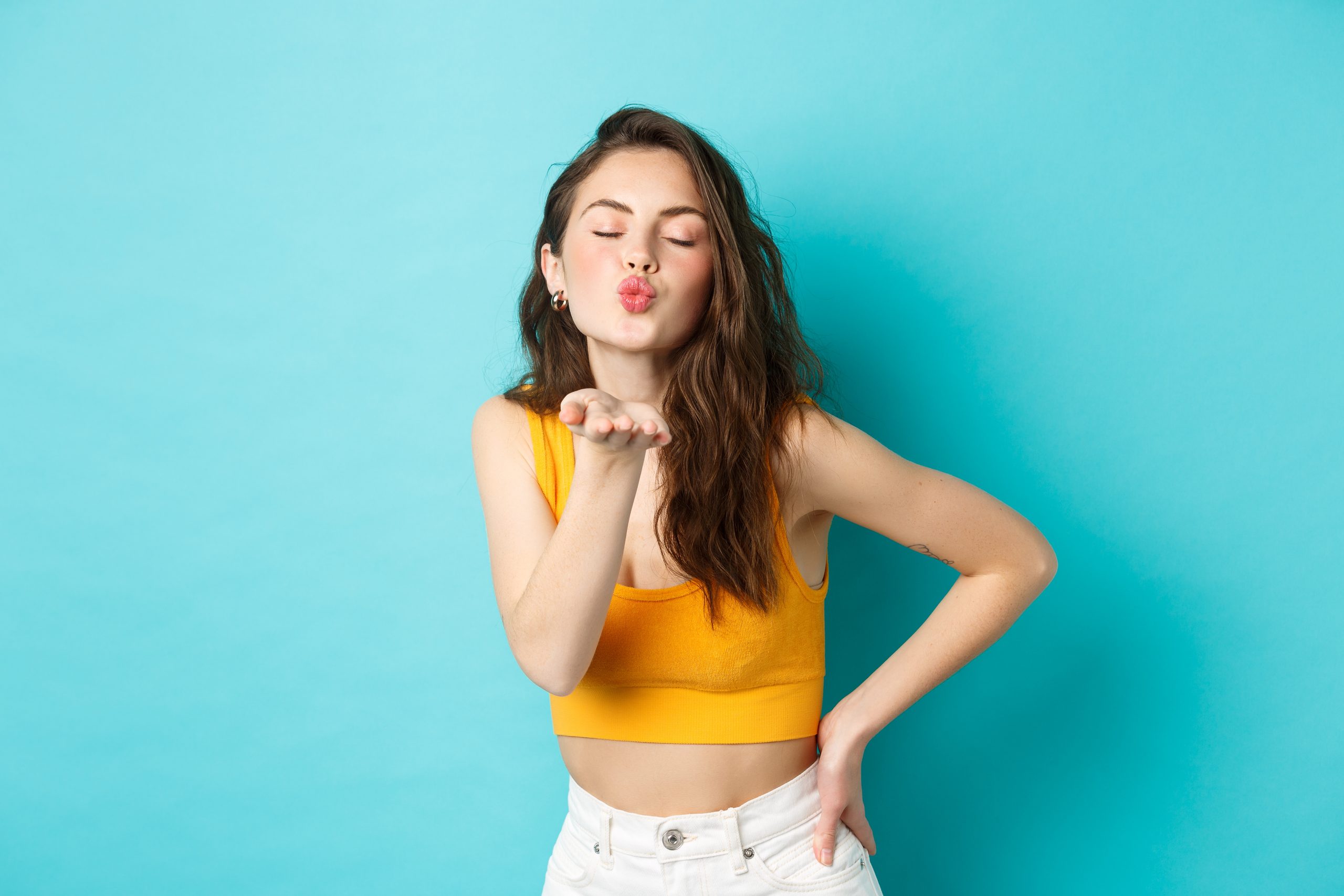 Coquettisches Sommermädchen, das einen Luftkuss auf Kamera, Kissen auf Lippen und nahe Augen schickt, steht auf blauem Hintergrund