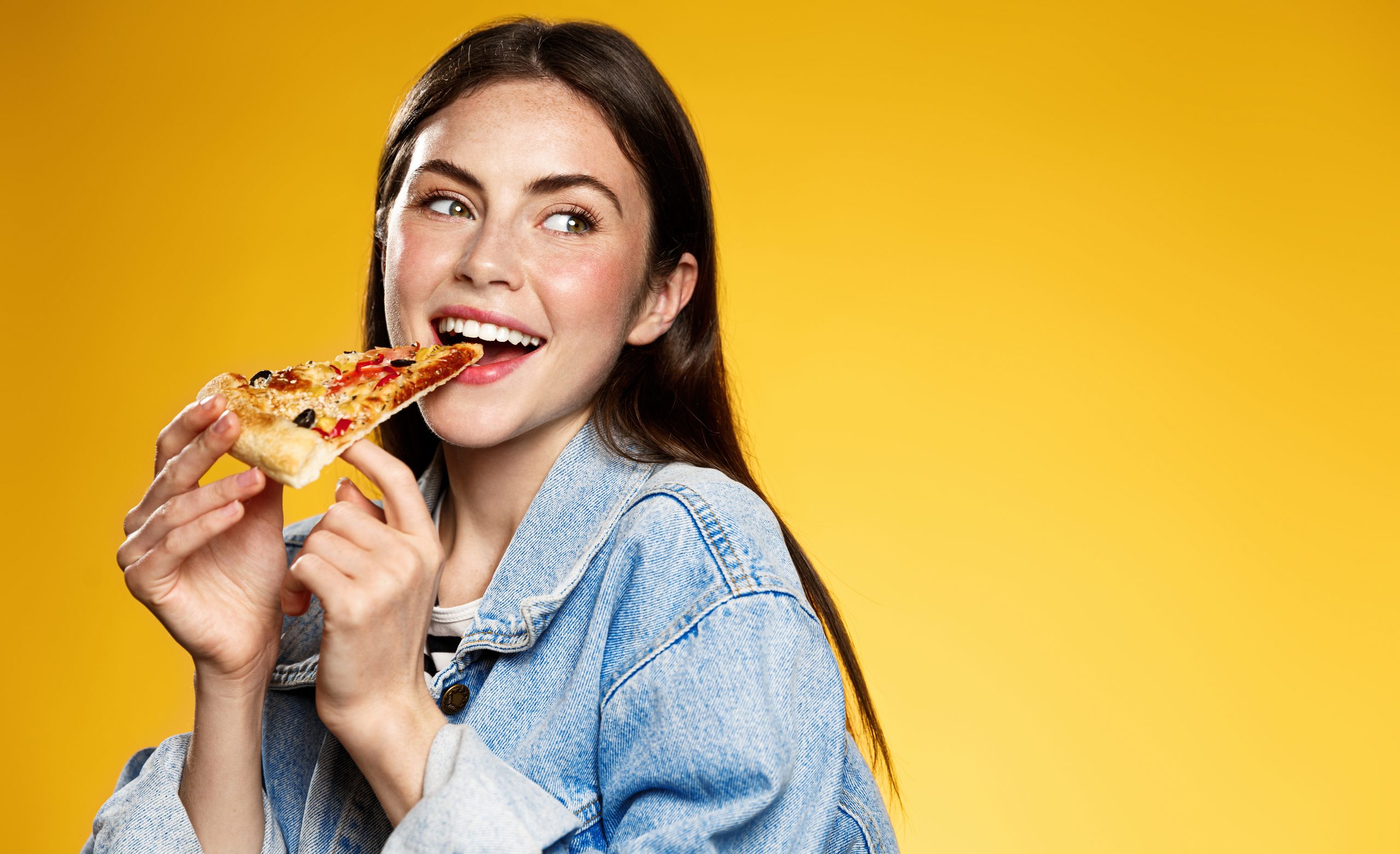 Foto von attraktiven Frauen isst eine Scheibe Pizza, Punkte neben Daumen, gekleidet in modischen Kleidung, Shows, wo Pizzeria ist, einzeln auf gelbem Hintergrund. Hübsches Mädchen hat Snacks mit Fastfood