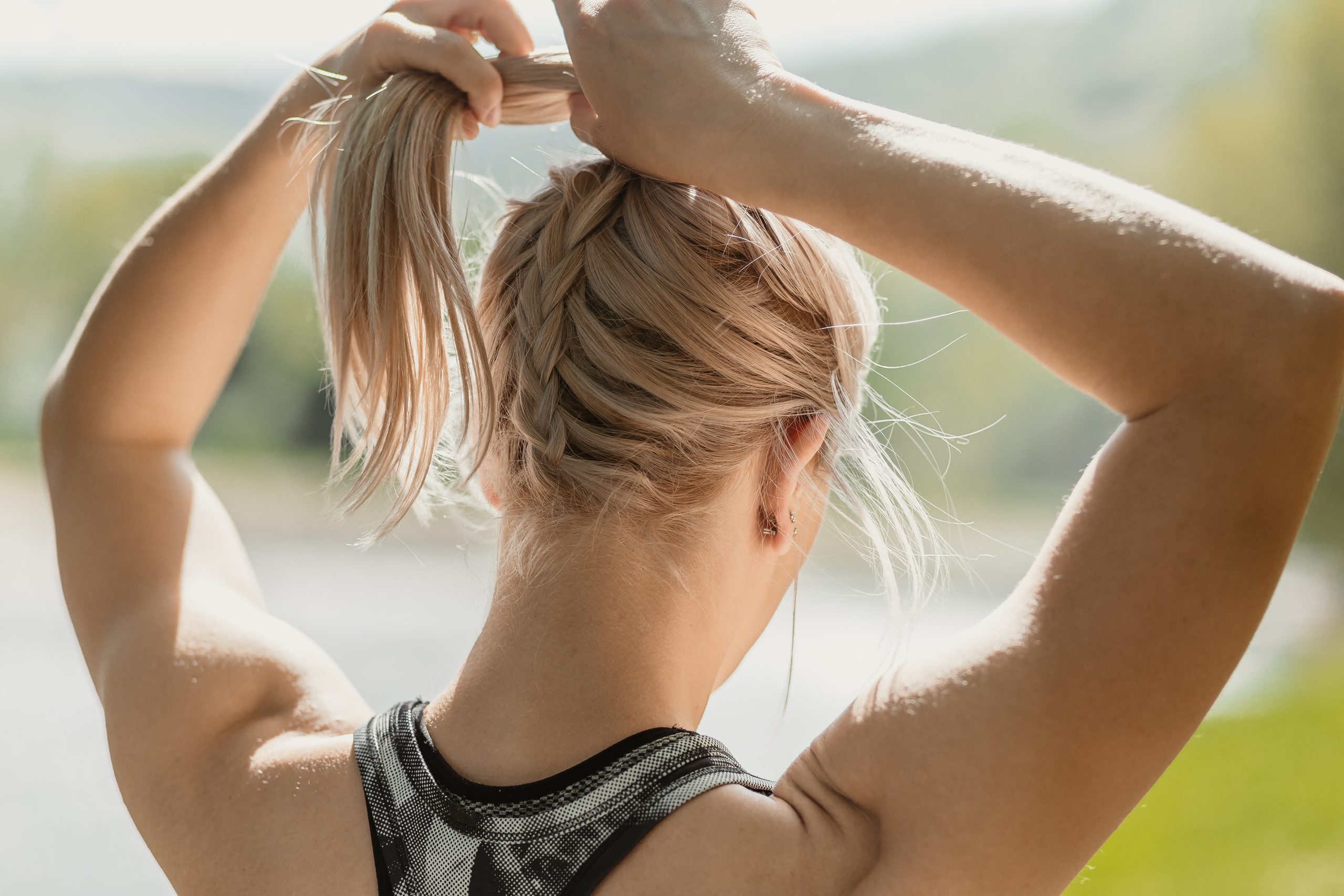 Back view photo of young blonde girl wearing sportswear tying a ponytail in the nature, by the river in the morning. Sunny weater. Healthy lifestyle and sport concept
