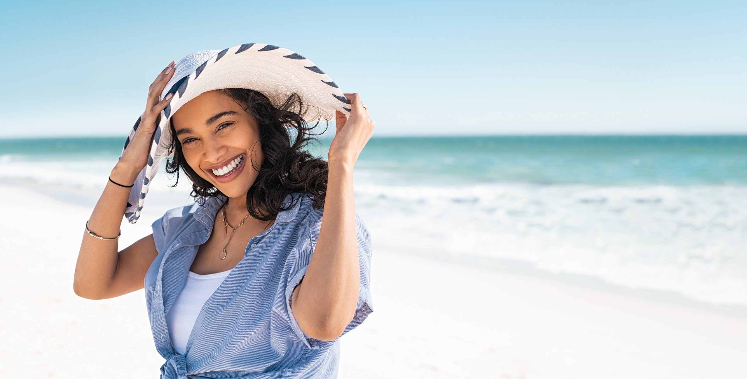Portrait von stylischer Latinenhispanie mit weißem Strohhut am Strand. Junge Lächelnde im Urlaub genießen Meeresbrise mit Strohhut und Blick auf die Kamera. Attraktives schönes Mädchen.
