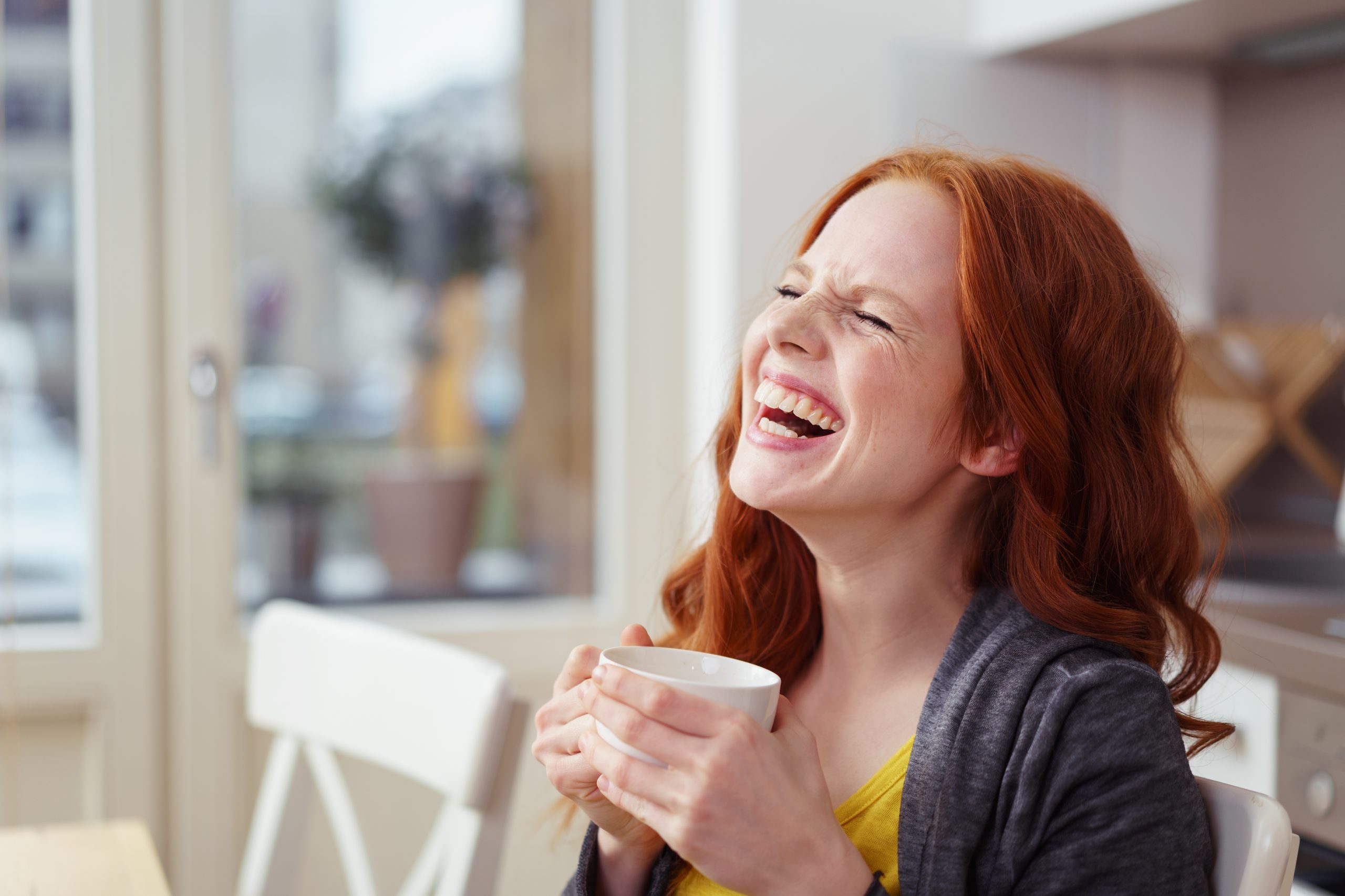 Spontane attraktive junge Rotkopffrau, die zu Hause in der Wohnung gut über eine morgendliche Tasse Kaffee lacht