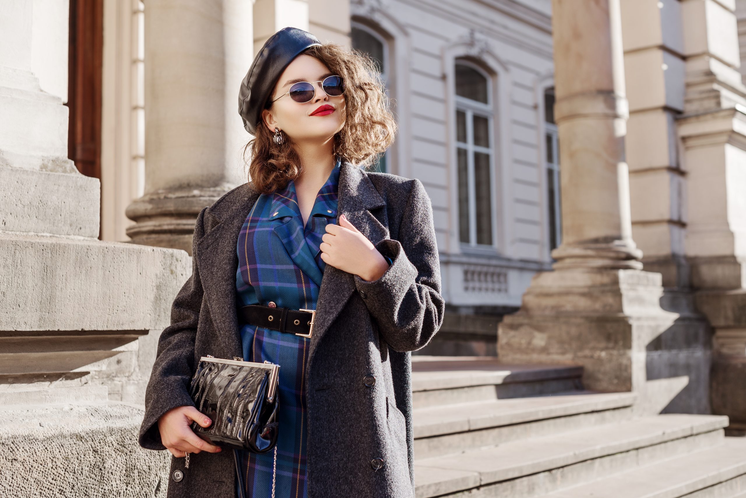 Outdoor Modeporträt von jungen schönen modischen Mädchen mit blauem Schachkleid, grauem Mantel, Lederbeet, Sonnenbrille, halten kleine Tasche, posieren in der europäischen Stadt. Kopieren, Leerraum