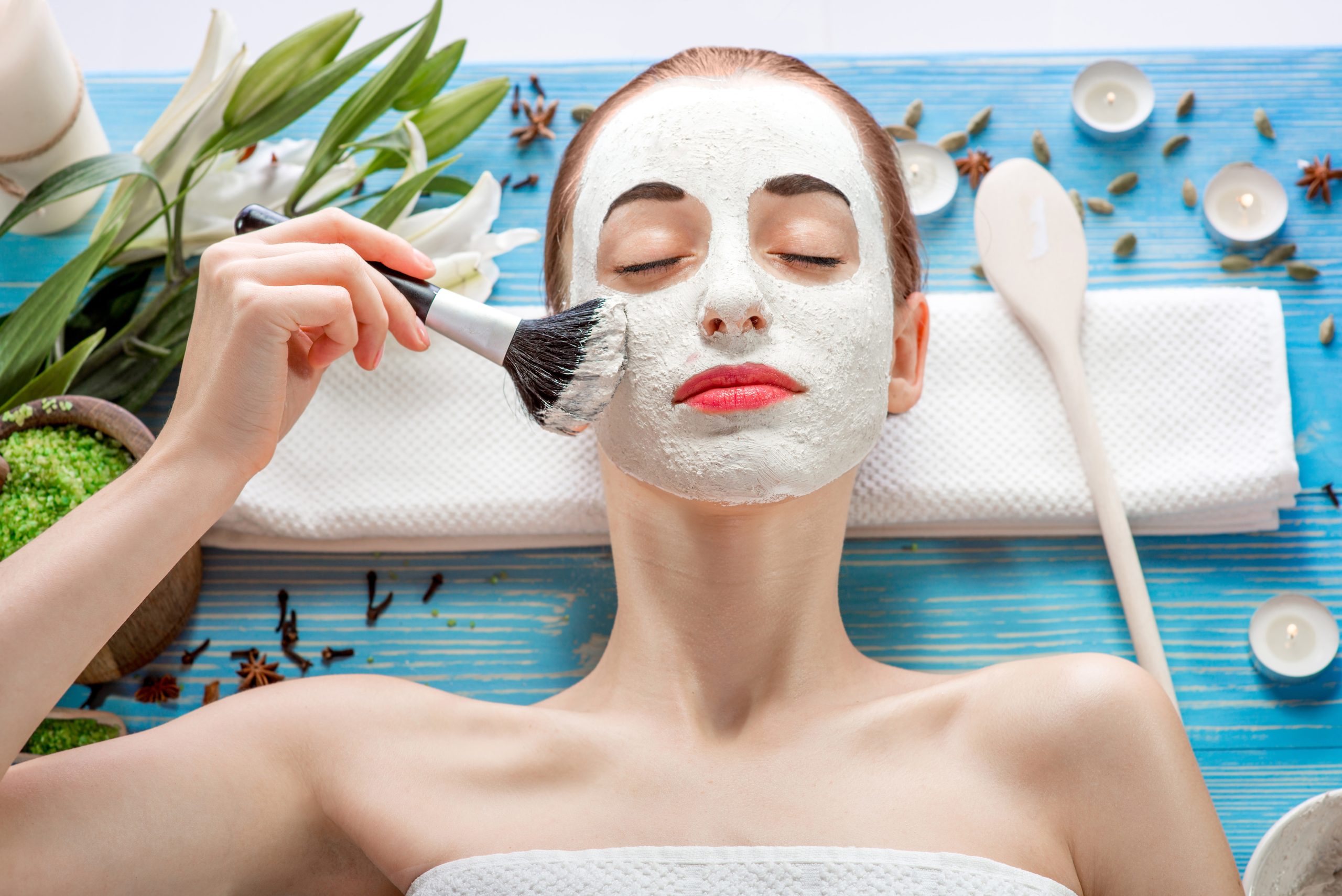 Young woman with spa facial mask on her face lying on blue table with flower, candles and sea salt in the beaty salon