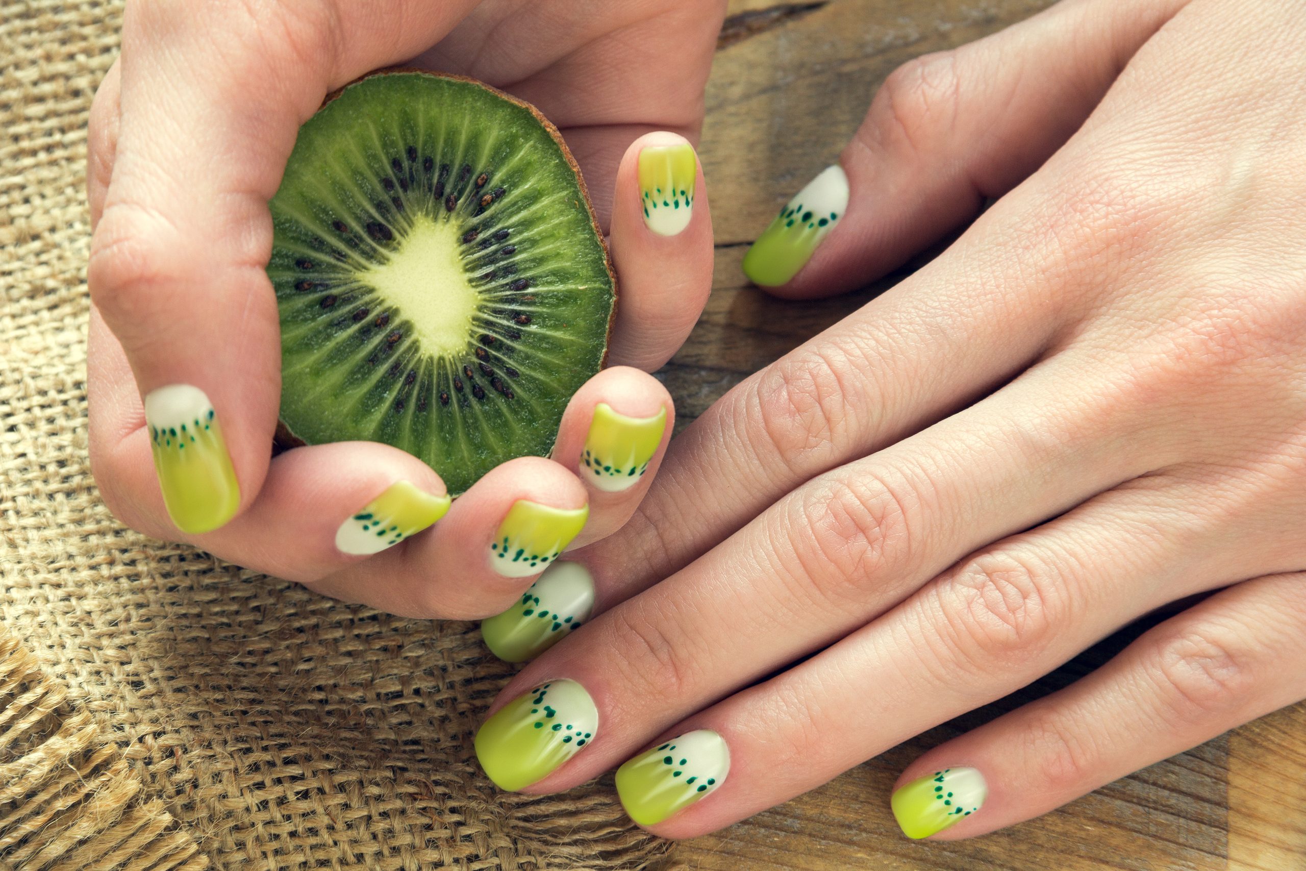 Kiwi and skin care of a beauty female hands with green and white moon nail art manicure on a sackcloth and wooden background