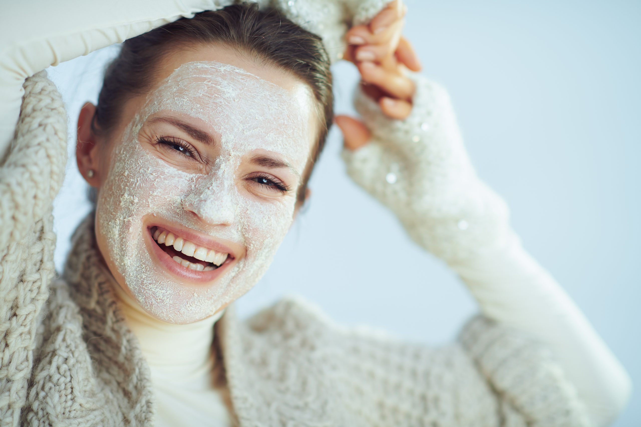 Portrait of smiling modern woman in roll neck sweater and cardigan with white facial mask as part of winter skin care isolated on winter light blue background.