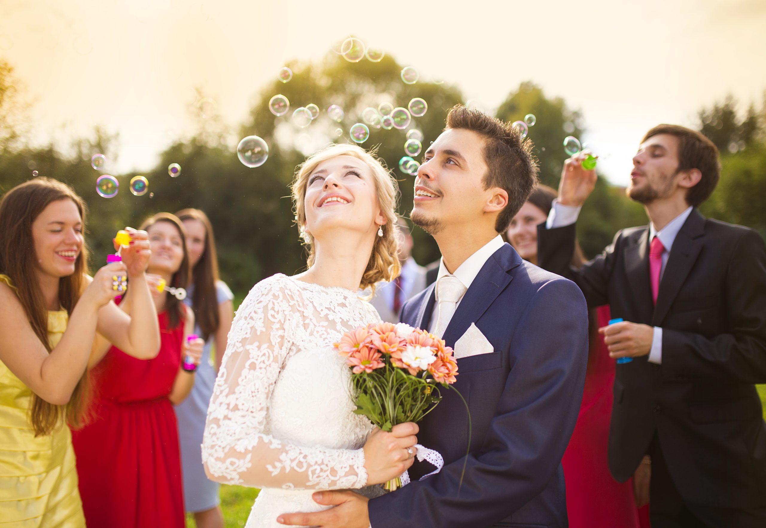 Young newlyweds enjoying romantic moment together at wedding reception outside, wedding guests in background blowing bubbles