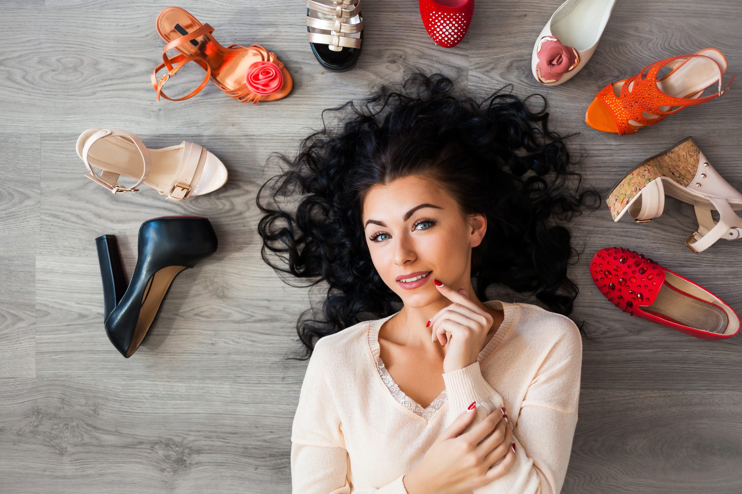 Beautiful young brunette cute funny girl lying on the floor around her laid out different shoes, sandals, ballet flats, a girl can not choose what kind of shoes to wear, fashion, emotion, top view