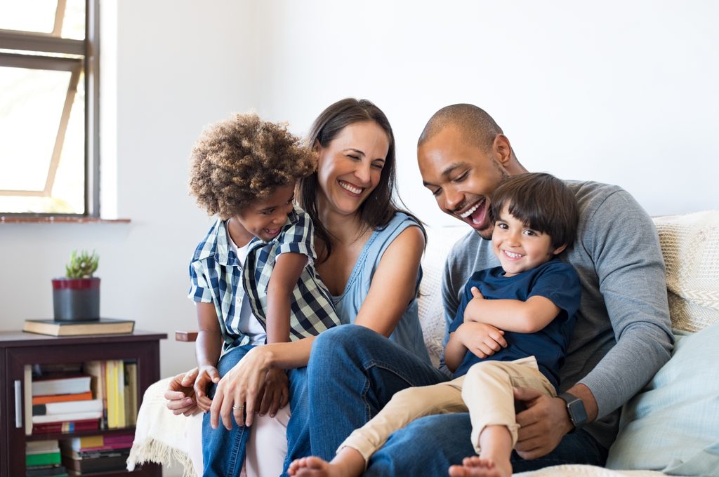Fröhliche, multiethnische Familie, die auf einem Sofa sitzt und zusammen lacht. Fröhliche Eltern, die zu Hause mit ihren Söhnen spielen. Schwarzer Vater kitzelt seinen kleinen Jungen, während Mutter und Bruder lächeln.