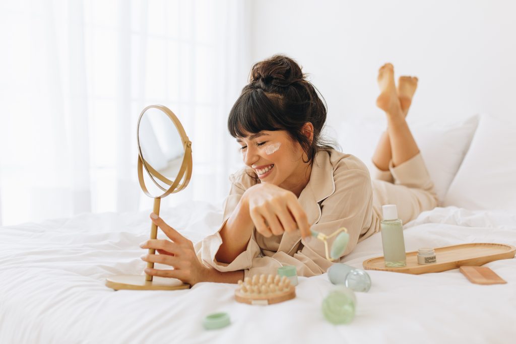 Nahaufnahme einer lächelnden jungen Frau, die auf dem Bett liegt und Gesichtscreme aufträgt. Frauen, die Schönheitspflege machen Gesichtsmassage zu Hause.