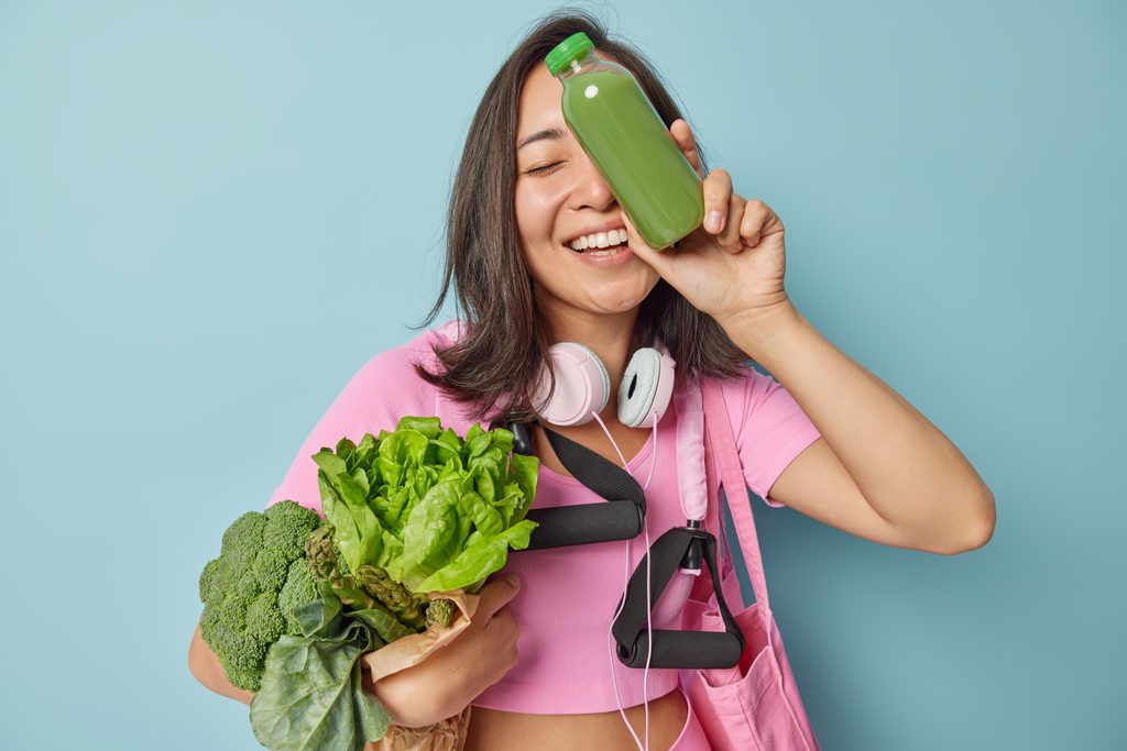 Happy Frau mit dunklem Haar hält eine Glasflasche frischer Smoothie tragen Gemüse Sport-Ausrüstung in Sportbekleidung gekleidet führt gesunden Lebensstil einzeln auf blauem Hintergrund. Geeignete Ernährung