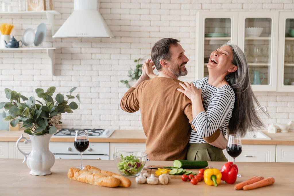 Fröhliche Eltern im mittleren Alter, die zusammen in der Küche tanzen, bereiten Essen für ein romantisches Abendessen zu, verbringen Zeit zusammen. Aktive Senioren